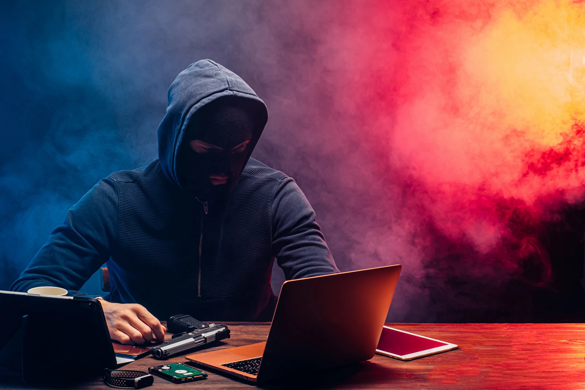 Vermummte Person mit Kapuzenpullover vor einem Laptop in rot-blauem Licht mit Festplatte und technischem Equipment